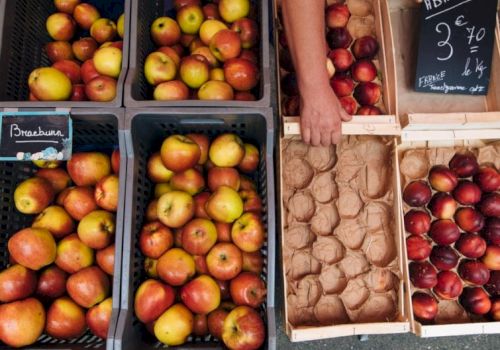 A market display of apples in crates and a rack of eggs and fruit, with a hand reaching for apples, and a chalkboard sign nearby, pretty and colorful.