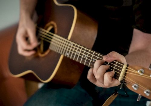 A person playing an acoustic guitar, fingers on the fretboard, strumming near the soundhole, close-up of hands and guitar neck.
