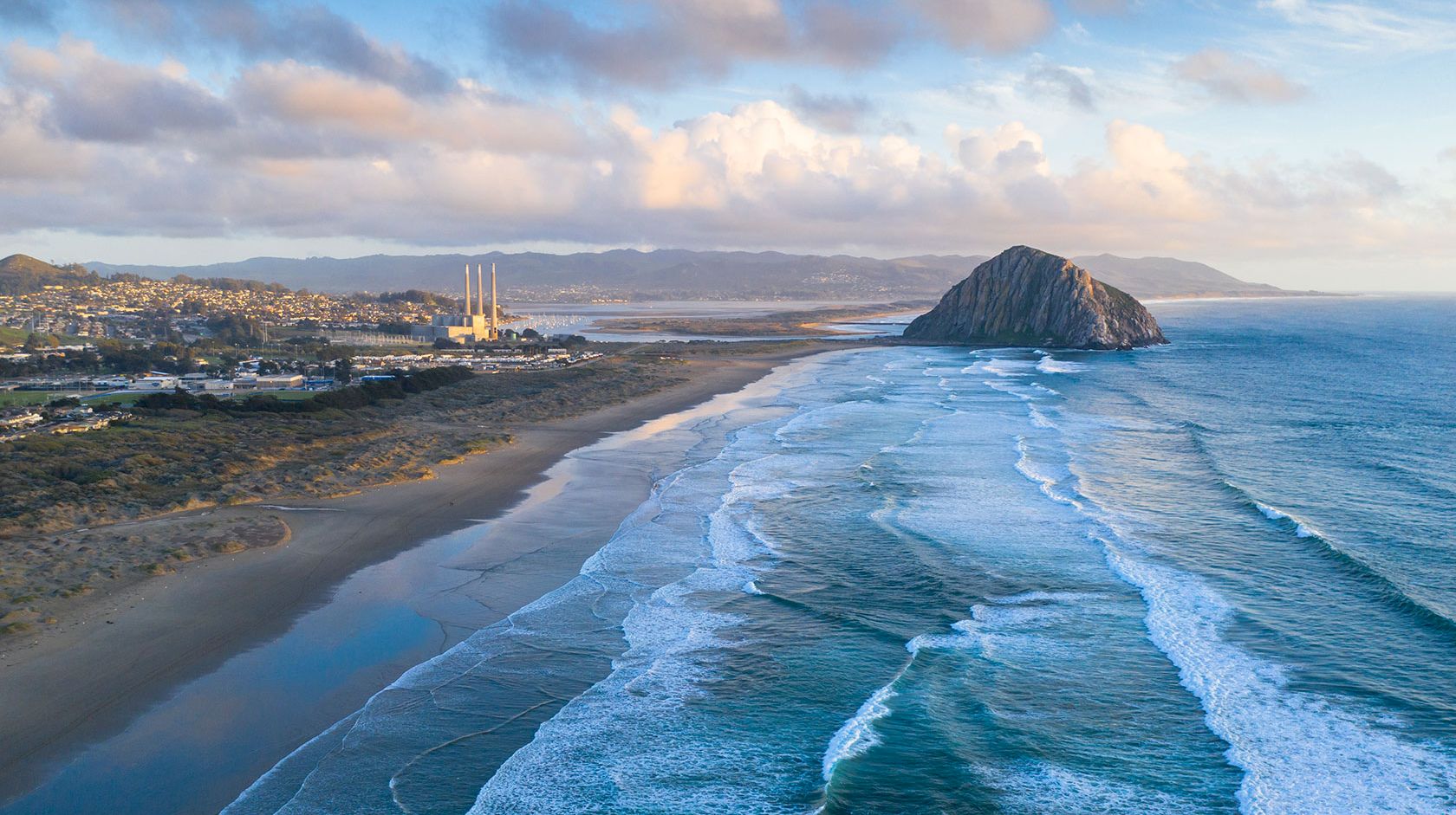 A coastal scene with a long beach, blue ocean waves, a distant town, and a large rocky islet off the shore, under a partly cloudy sky.
