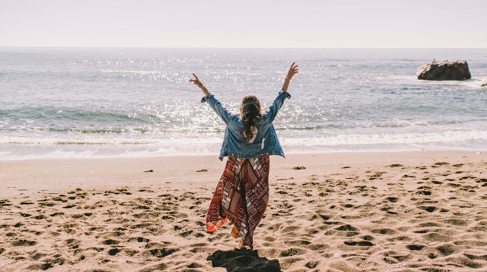 A person in a flowy skirt and denim jacket dances on a sunny beach, arms raised, waves shimmering in the background.