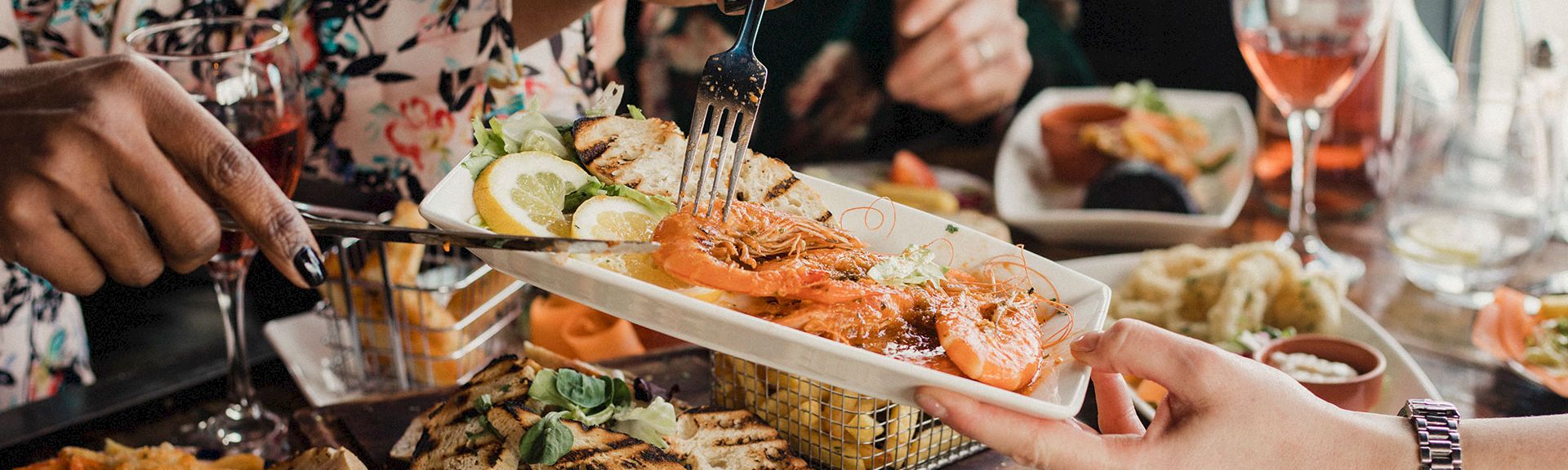 People sharing a seafood platter at a lively table, cutting into shrimp with lemons, wine glasses, and assorted side dishes.