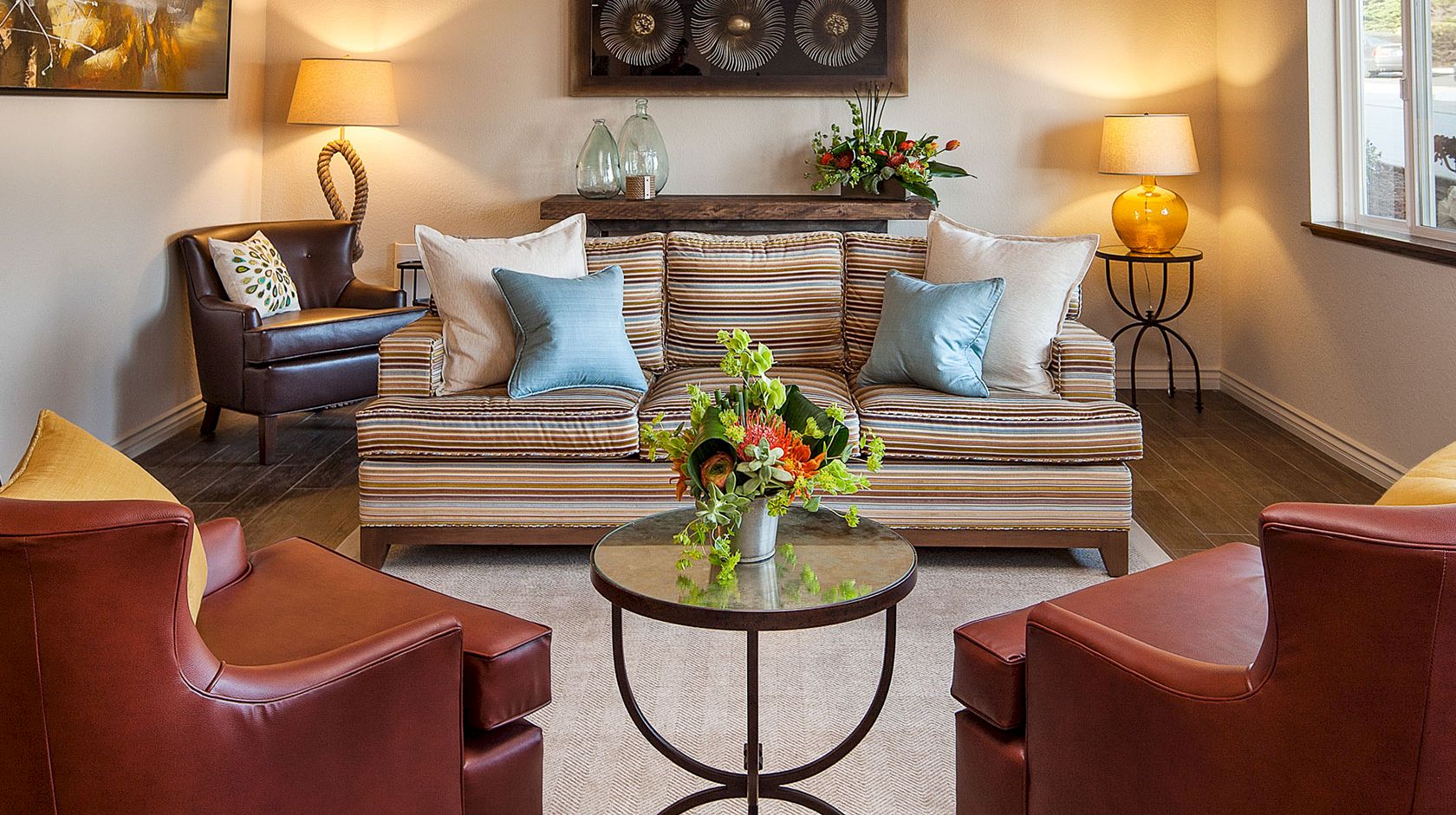 A cozy living room with striped sofa, colorful cushions, two red armchairs, a glass-top coffee table, and warm lamps creating a welcoming vibe.