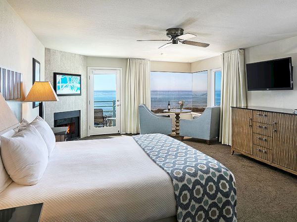 A bright hotel room with a king bed, fireplace, flat-screen TV, and a balcony overlooking the ocean, perfect for a relaxing coastal stay.