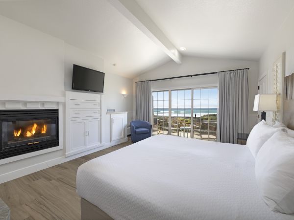 A bright hotel room with a white-trimmed fireplace, built-in cabinet, wall-mounted TV, a king bed, a blue chair, and a sliding door to a balcony overlooking the ocean, ending this sentence.