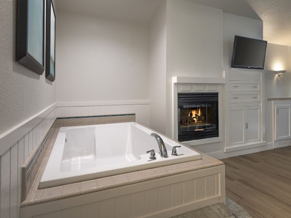 A modern bathroom with a built-in soaking tub, a gas fireplace, white cabinetry, and a wall-mounted TV in a cozy, light-filled space.