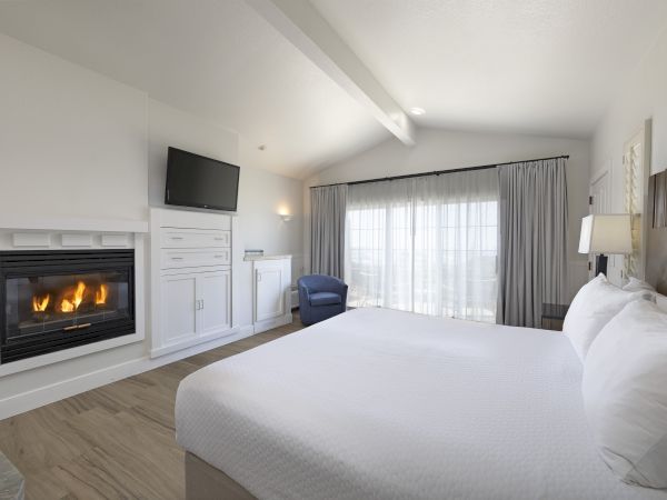 A bright hotel room with a lit fireplace, mounted TV, white cabinetry, a large bed, blue armchair, and sliding glass doors with sheer curtains.