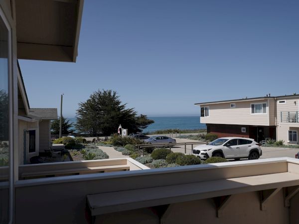 A sunny beach town scene: white coastal apartments, parked cars, a clear blue sky, and the ocean in the distance beyond a small garden.
