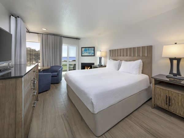 A bright hotel room with a queen bed, two nightstands, a flat-screen TV, a seating area, and a large window with a view of scenery.