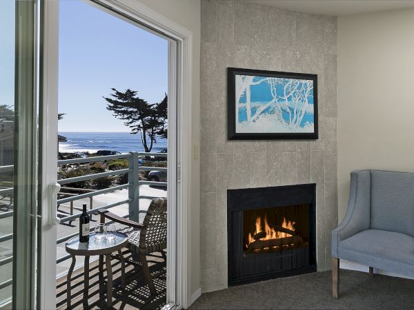 Cozy hotel room with a balcony view of the ocean, a lit fireplace, a light blue chair, and a framed artwork above the fireplace.