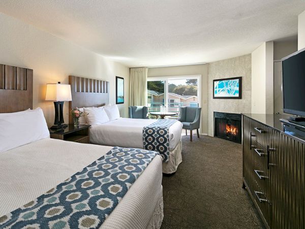 Two queen beds in a modern hotel room with a fireplace, flat-screen TV, and a seating area by a window.