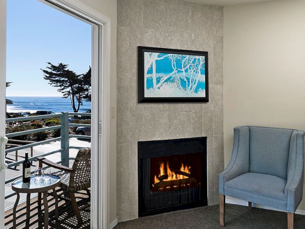 Cozy hotel room with a lit fireplace, a blue armchair, a framed ocean-themed painting, and a balcony view of the sea and trees outside.
