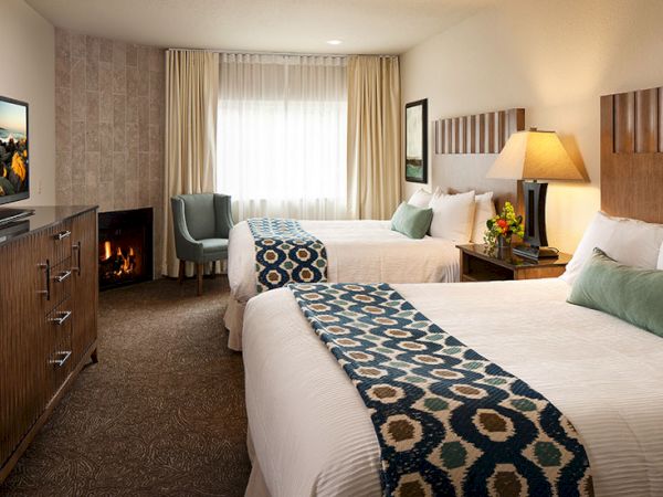 A cozy hotel room with two neatly made beds, a patterned runner, a desk and TV, a lamp, and a window with sheer curtains by a seating area.