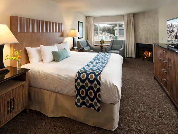 A cozy hotel room with a king bed, geometric throw, side tables with lamps, a seating area by the window, and a fireplace by the dresser.