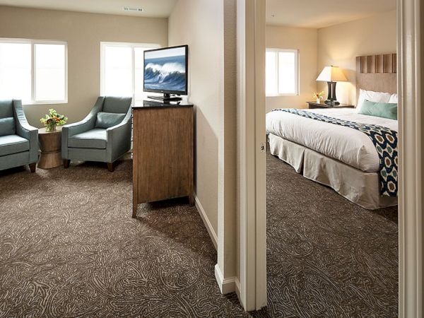 Hotel suite with a living area and bedroom separated by a doorway; two blue chairs, a small table, and a TV in the living space. End.