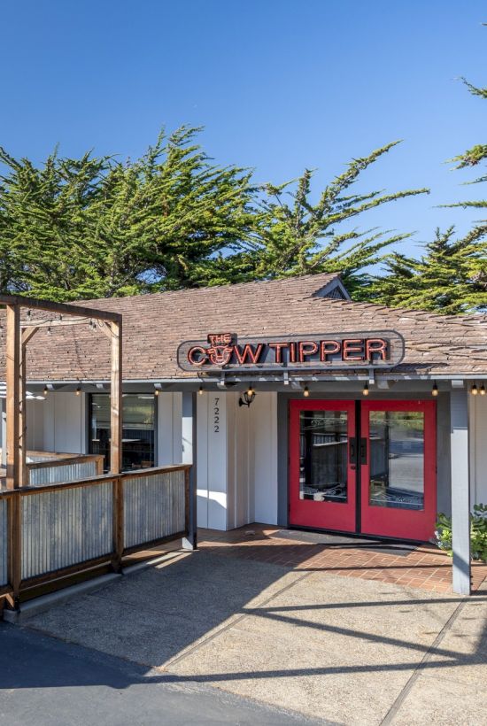 A small outdoor cafe or restaurant with a wooden fence enclosure, patio seating, red doors, and a sign that reads &ldquo;Wipper&rdquo; on a sunny day.