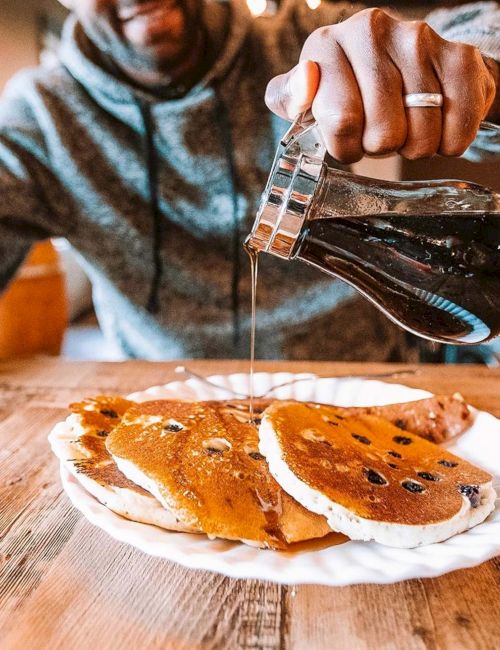 Someone pours syrup over chocolate chip pancakes on a wooden table at a cafe, a casual morning scene.