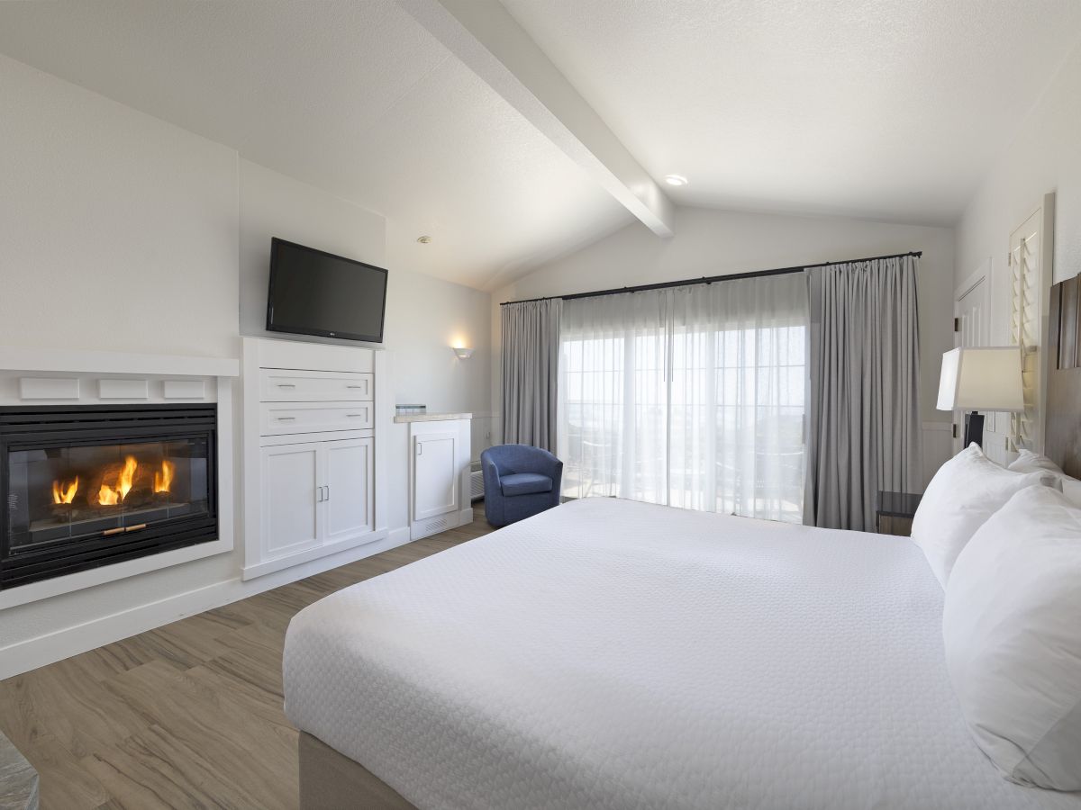 A bright hotel room with a lit fireplace, wall-mounted TV, white cabinetry, a comfy blue chair, large window with curtains, and a neatly made white bed.