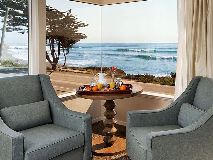 Cozy seaside lounge with two gray armchairs framing a round table, overlooking a sunny beach and ocean through large windows.